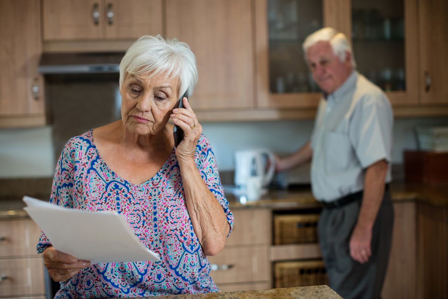 Oudere vrouw aan de telefoon met een briefje in haar hand, oudere man op de achtergrond