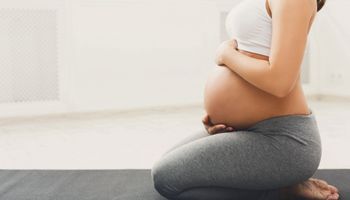 Mulher grávida irreconhecível praticando yoga em pose de herói, acariciando sua barriga. A jovem e feliz expectativa relaxa, pensa no seu bebé e no seu futuro.