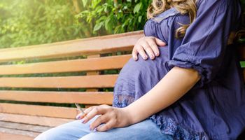Een zwangere vrouw zit op een bankje in het park en rookt een sigaret.