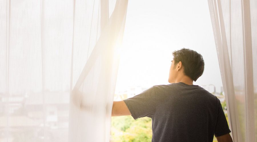 One person pushes aside a light window curtain to allow the sun to shine through.