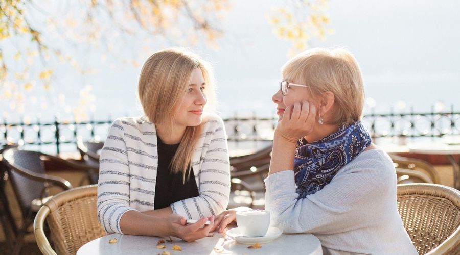 Uma jovem mulher senta-se num café lá fora com a mãe e olham-se uma para a outra. As montanhas podem ser vistas em segundo plano.