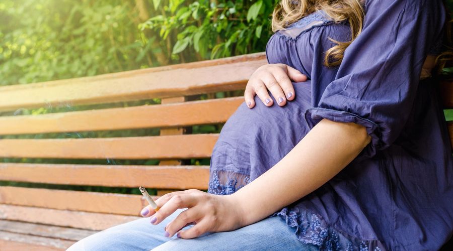 A pregnant woman sits on a park bench smoking a cigarette.