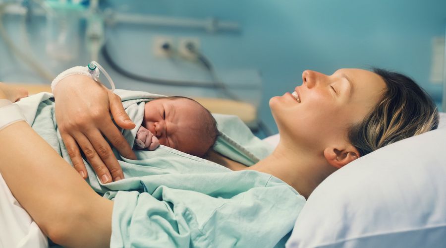 Eine Mutter haltet liegend ihr neugeborenes Kind auf ihrer Brust in einem Krankenhaus und lächelt dabei mit geschlossenen Augen.
