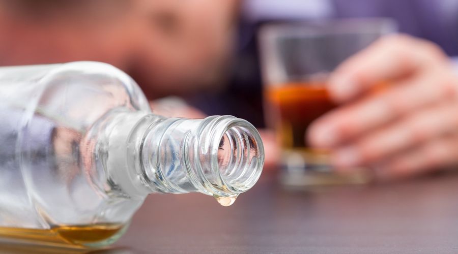 Grande plano de uma garrafa de whisky caída, quase vazia, com uma pessoa a dormir com um copo de whisky meio cheio no fundo.