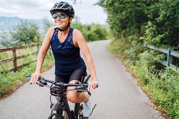 Portrait of a happy smiling woman dressed in cycling clothes, helmet and sunglasses riding a bicycle on the asphalt out-of-town bicycle path. Active sporty people concept image.