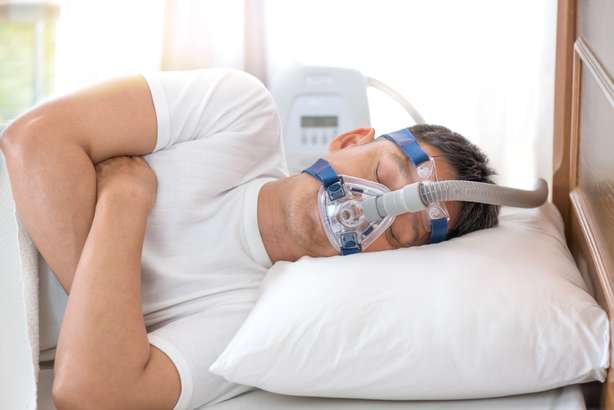 Un señor mayor lleva una máscara respiratoria mientras está tumbado de lado en la cama intentando conciliar el sueño.