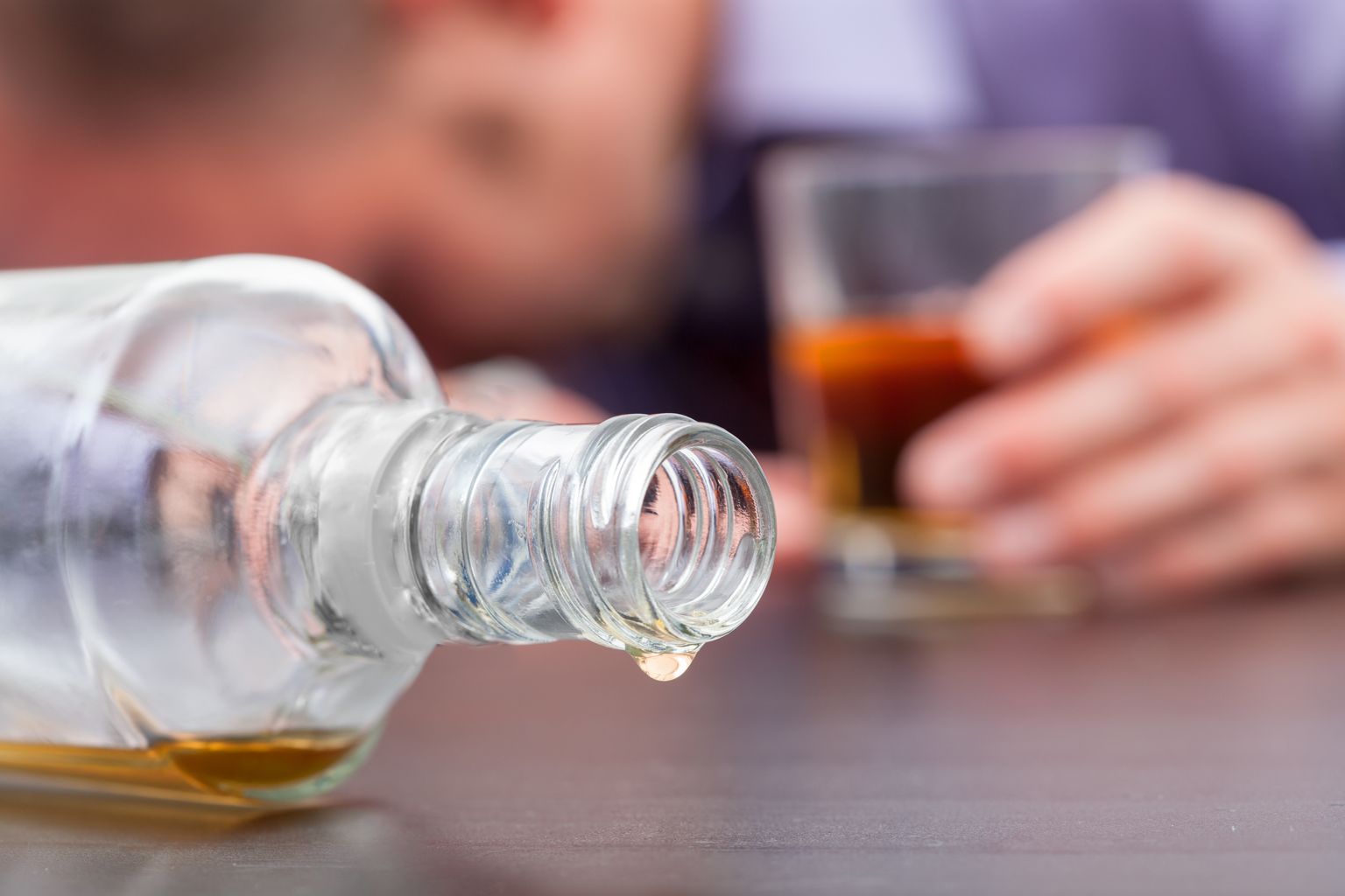 Gros plan sur une bouteille de whisky renversée et presque vide avec, en arrière-plan, une personne endormie avec un verre de whisky à moitié plein.