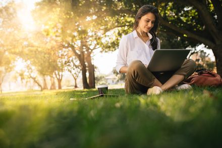 Frau sitzt auf Gras im Park und arbeitet auf Laptop. Frauen tragen Kopfhörer mit Laptop, während sie im Park unter einem Baum sitzen, mit hellem Sonnenlicht von hinten.
