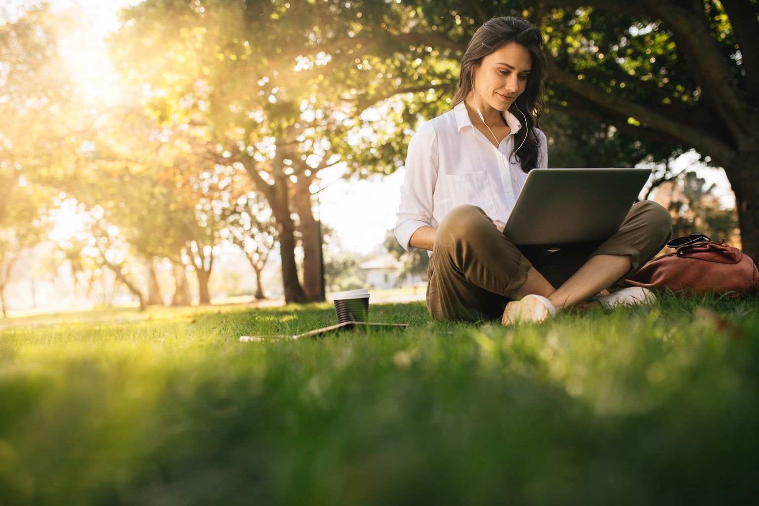 Vrouw zittend op gras in park werkend aan laptop. Vrouwen die een hoofdtelefoon met laptop dragen terwijl ze onder een boom in een park zitten met fel zonlicht van achteren.