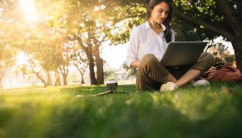 Woman sitting on grass in park working on laptop. Women wearing headphones with laptop while sitting under tree in park with bright sunlight from behind.