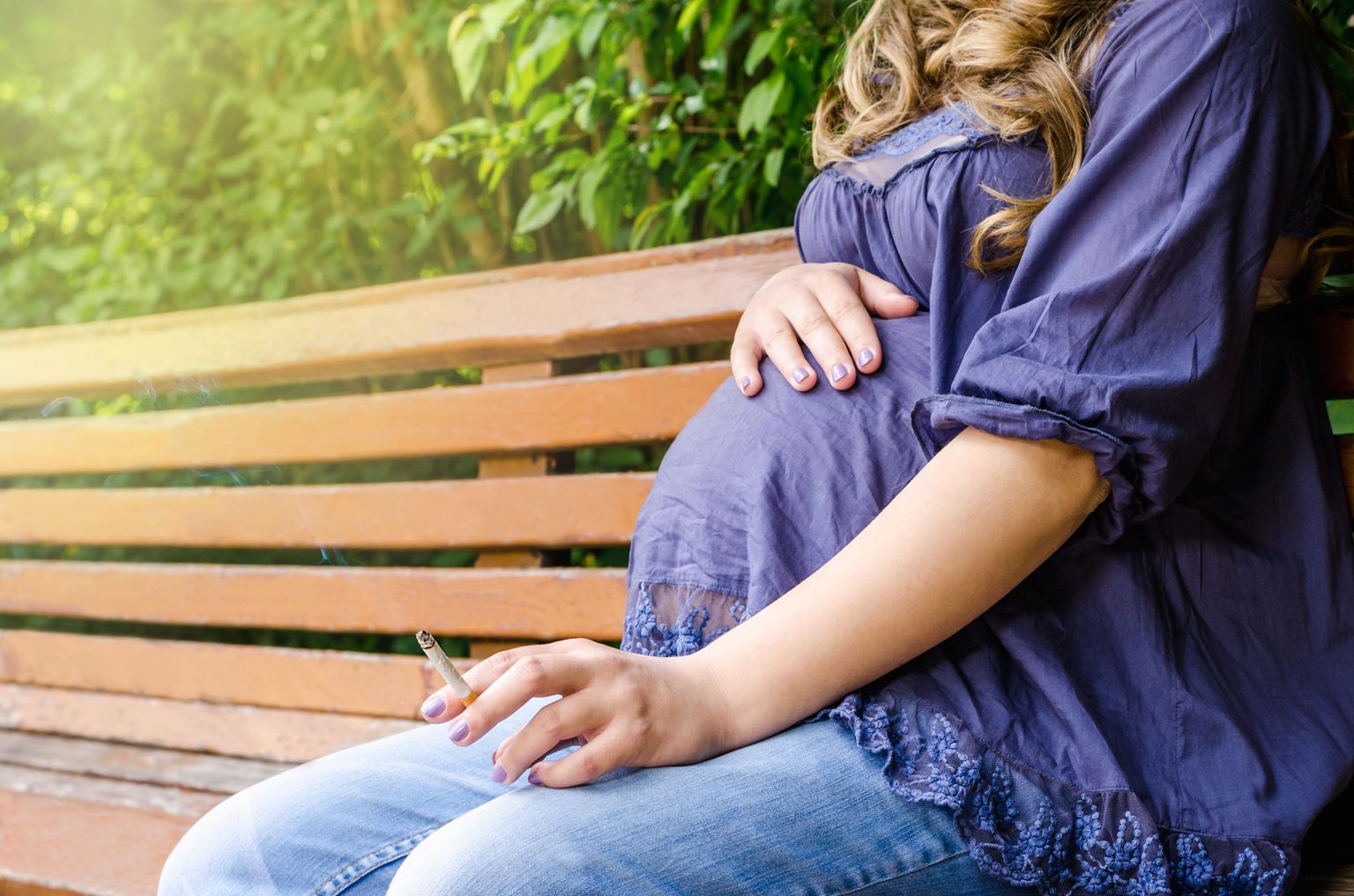 A pregnant woman sits on a park bench smoking a cigarette.