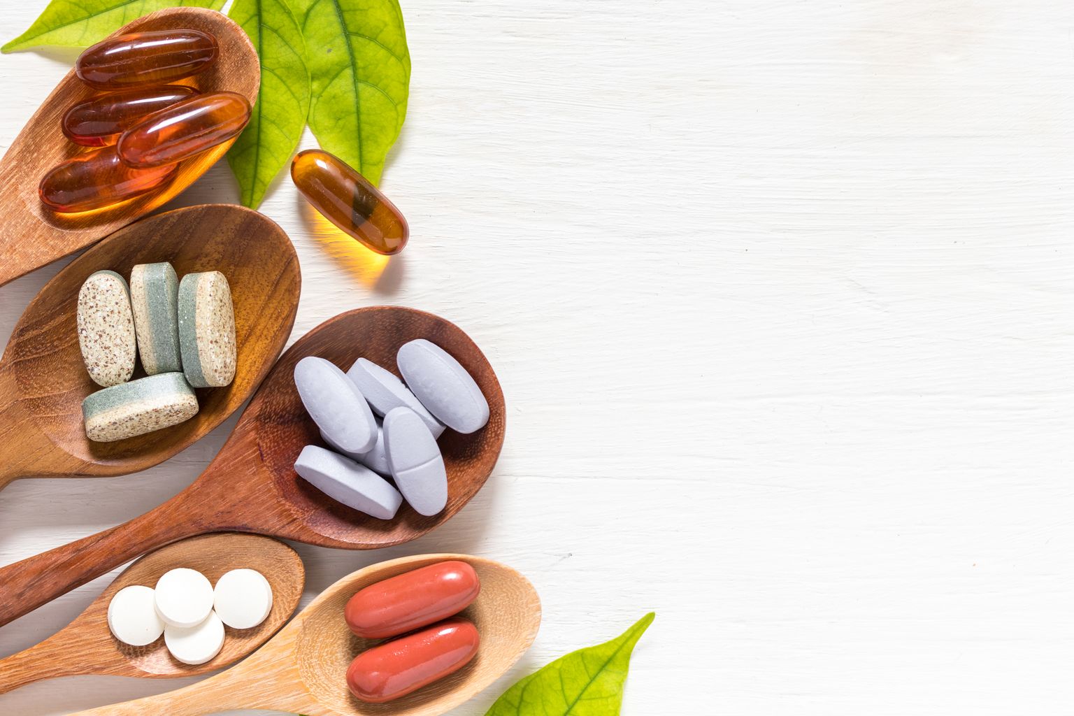 Variedade de comprimidos de vitaminas em colheres de madeira sobre fundo branco com folha verde, produto complementar e médico, superfície plana para leigos
