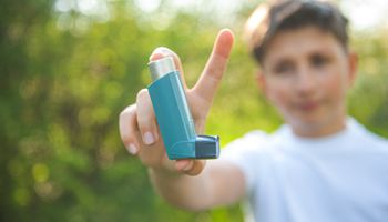 Een jongen in een wit T-shirt houdt een astma-inhalator in de camera. Op de achtergrond is zonnige natuur.