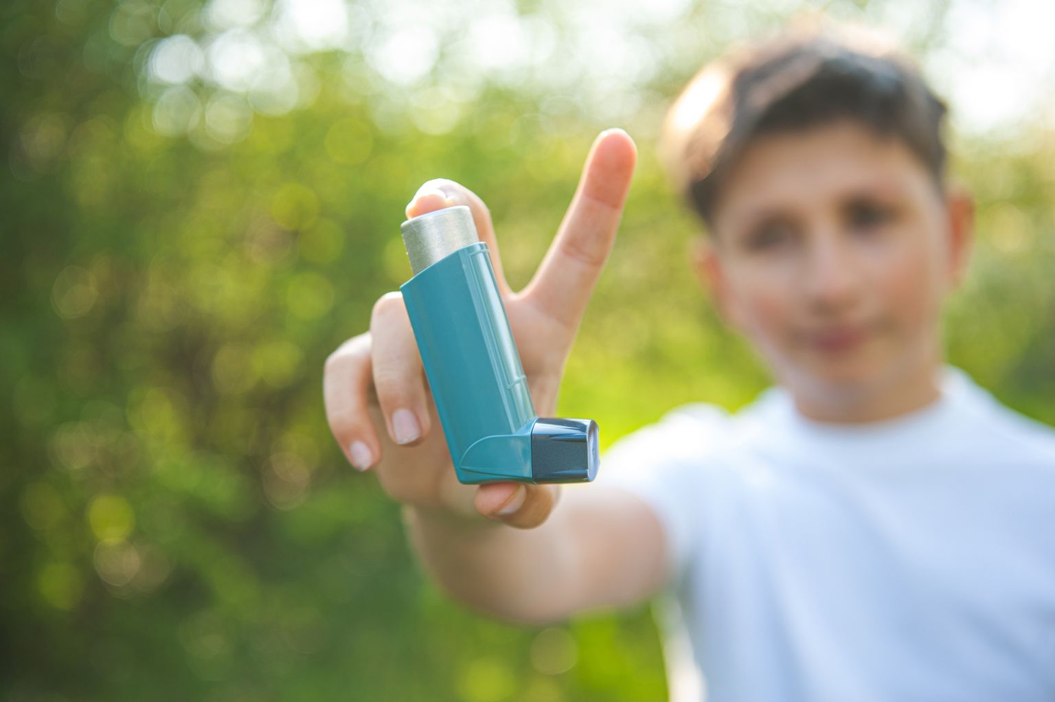 Een jongen in een wit T-shirt houdt een astma-inhalator in de camera. Op de achtergrond is zonnige natuur.