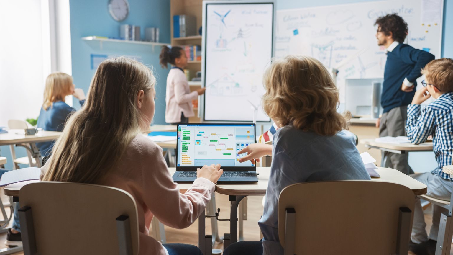 Classe de sciences à l'école primaire : Un petit garçon et une petite fille en bandoulière utilisent un ordinateur portable avec un écran montrant un logiciel de programmation. Un professeur de physique explique une leçon à une classe diversifiée d'enfants intelligents.