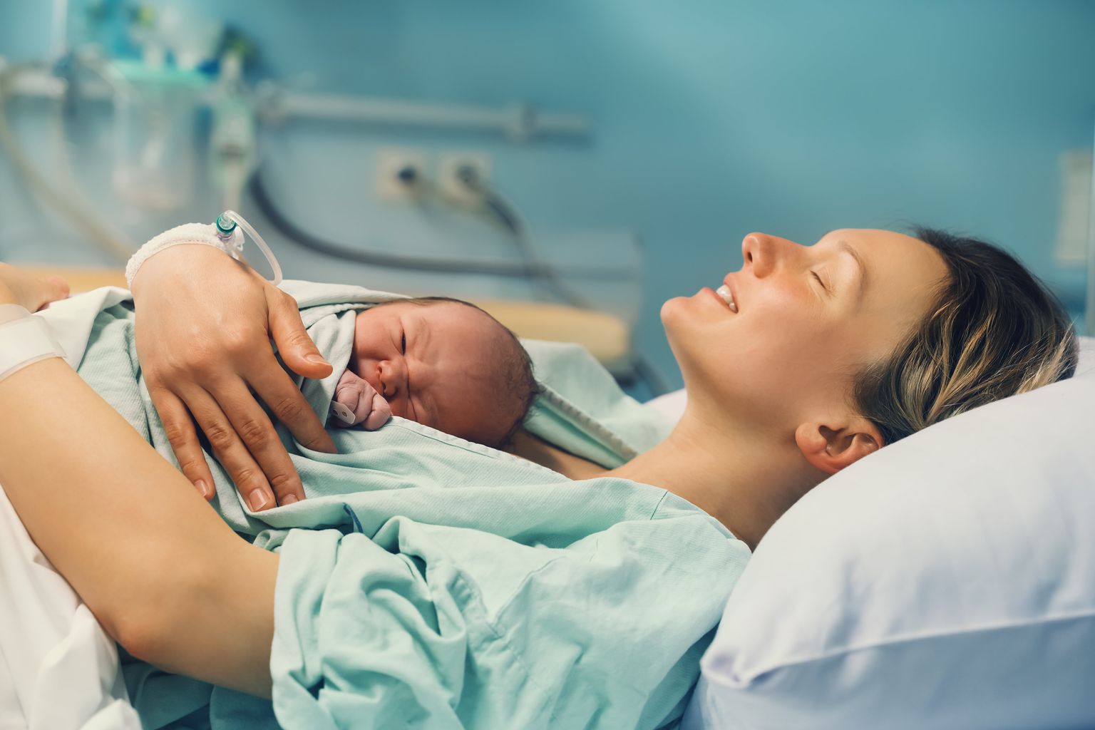 Uma mãe está deitada e segura o seu recém-nascido no peito num hospital, sorrindo de olhos fechados.