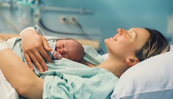 Een moeder ligt en houdt haar pasgeboren kind op haar borst in een ziekenhuis, glimlachend met haar ogen dicht.